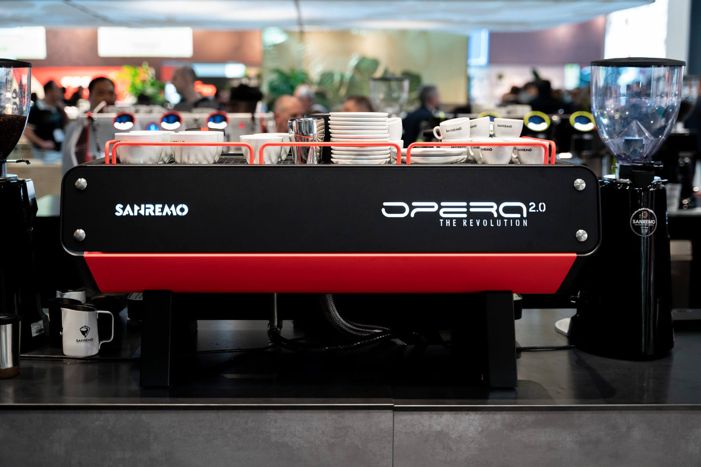 Sanremo booth at coffee equipment expo with crowd, showing a black and red Sanremo Opera Commercial Espresso Machine and a Coffee Grinder  from a rear view

